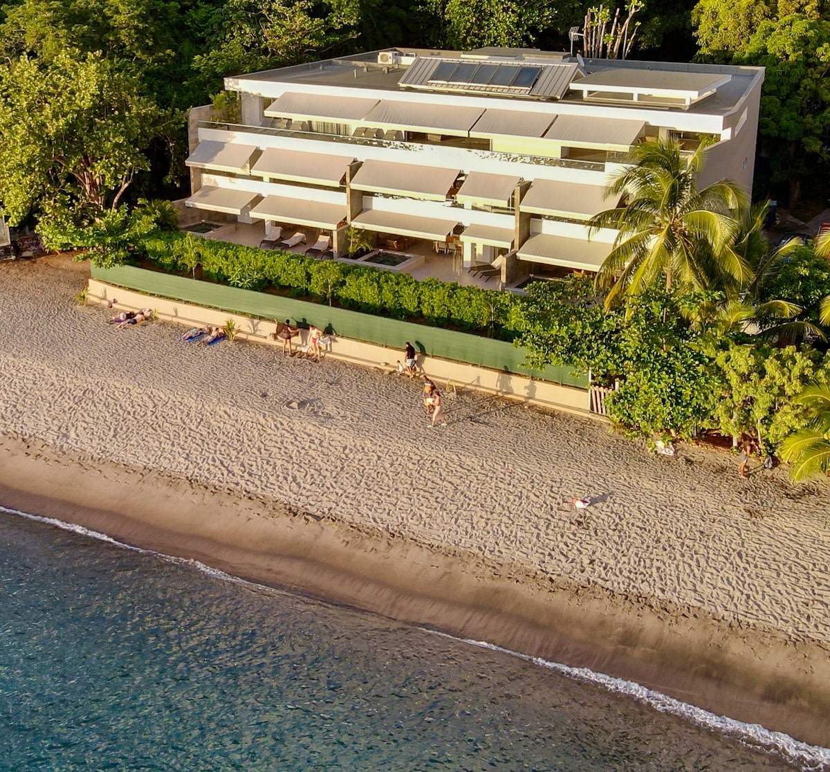 The apartment building is positioned along the beach, surrounded by lush greenery and palm trees. It features multiple terraces accessible from the upper levels, with a sandy beach appearing in the foreground where a few people can be seen enjoying the coast.