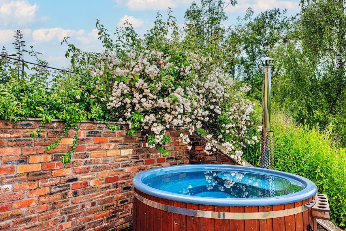 A wood-fired hot tub is set amidst lush greenery and blooming flowers, surrounded by a rustic brick wall. The clear blue water reflects the sky, creating a serene atmosphere ideal for relaxation outdoors.