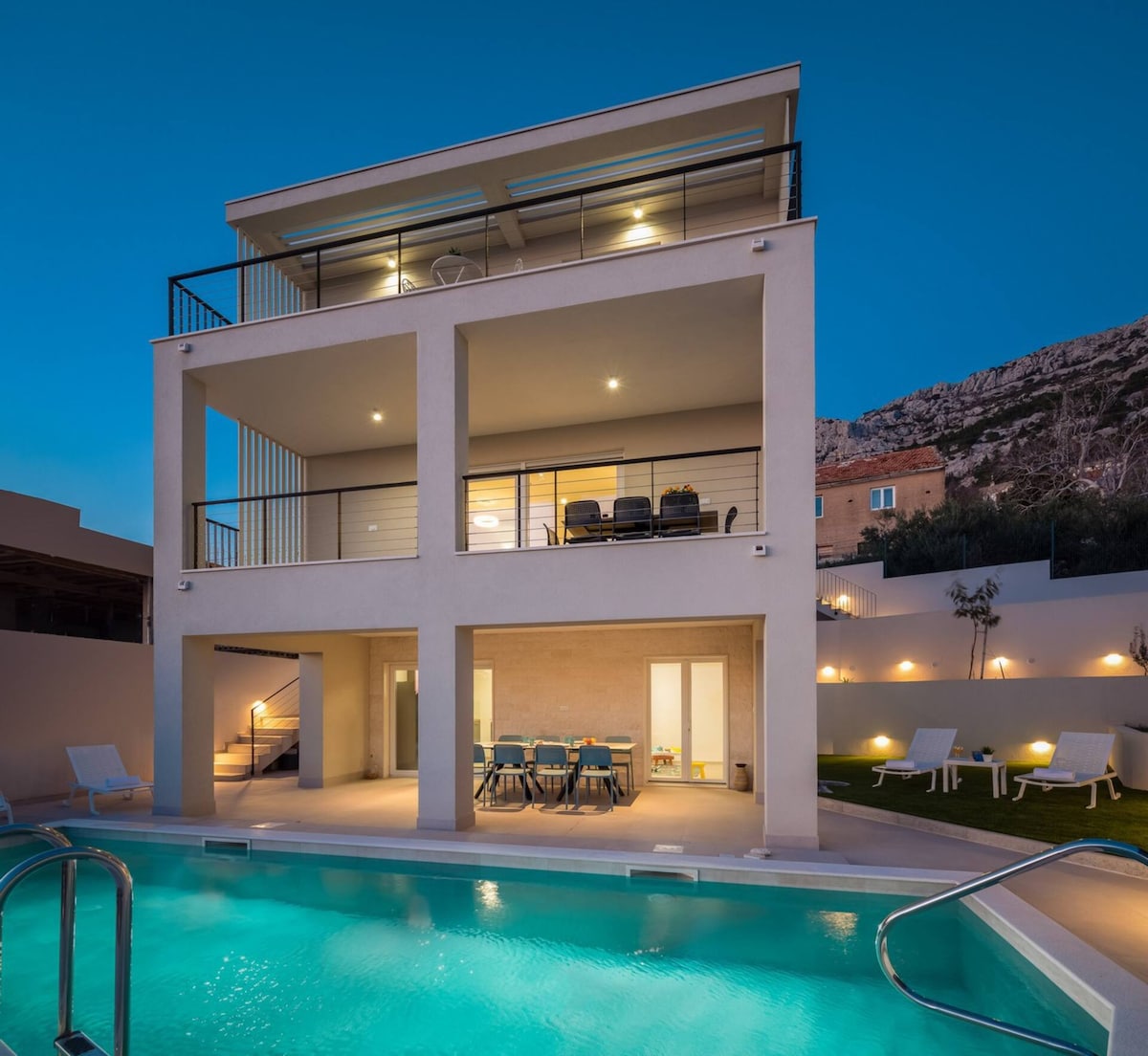 The modern villa showcases three levels, with a spacious outdoor pool at the forefront. Outdoor seating is visible on the terrace, and lounge chairs line the pool area. The exterior is illuminated, creating a serene atmosphere as dusk settles.