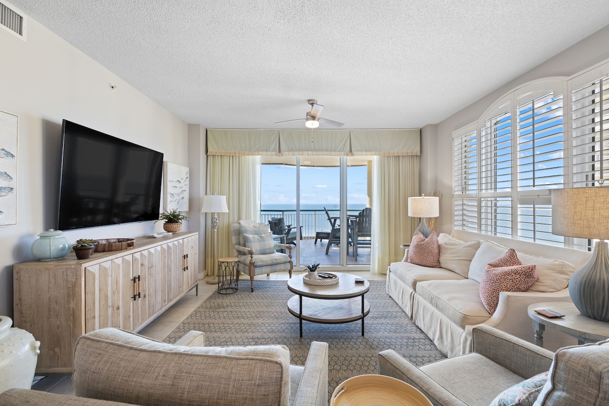 A spacious living area showcases designer furnishings, including light-colored sofas and accent chairs. A large television is mounted on a wooden console. The floor-to-ceiling windows allow natural light in and provide views of the private balcony and beachfront outside.