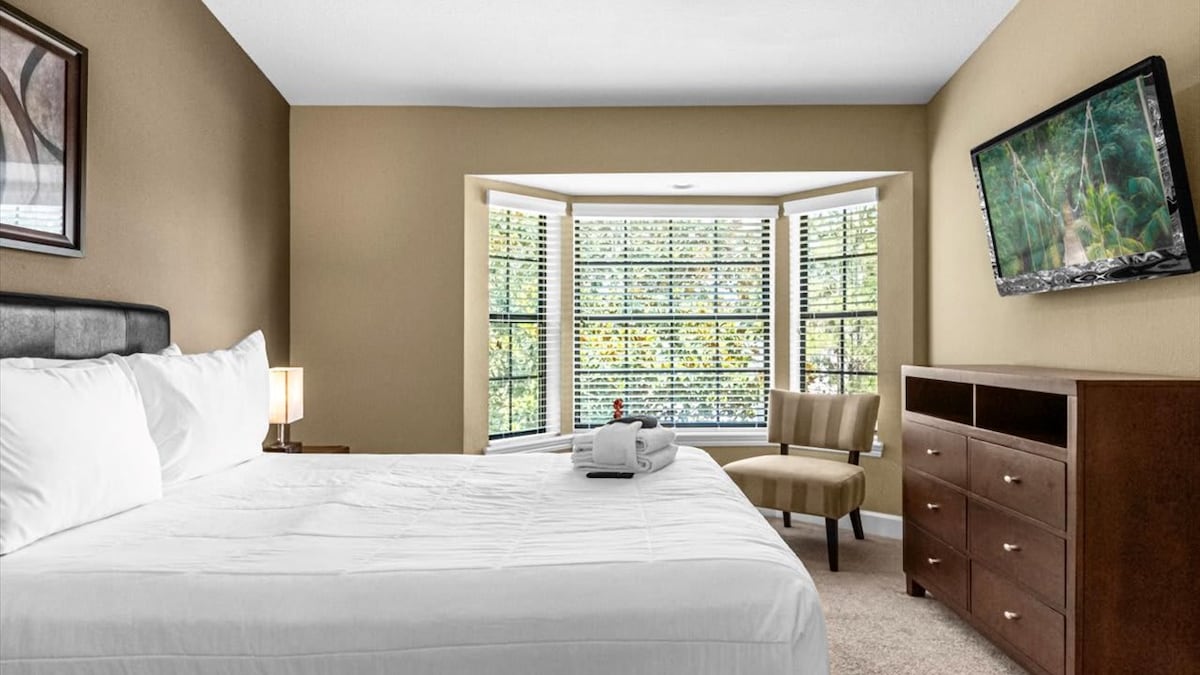 A well-appointed bedroom displays a spacious bed with crisp white linens. Natural light pours in through multiple bay windows, framed by lush greenery outside. A comfortable chair and a wooden dresser add functional elegance, while a flat-screen television is mounted on the wall.