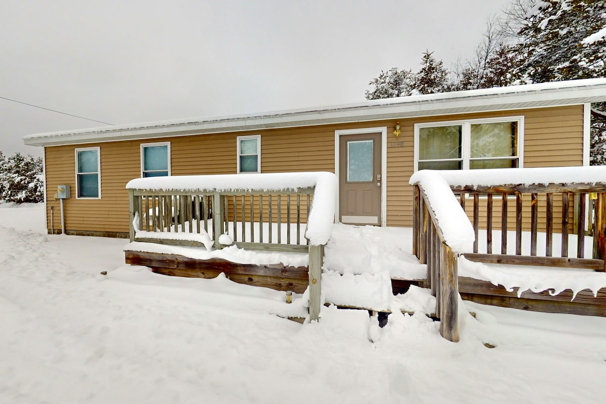 A single-story home is framed by a layer of fresh snow. The entrance features a front porch with wooden railings, partially covered in snow. Two windows flanking the door allow natural light to enter, while snow blankets the ground nearby.