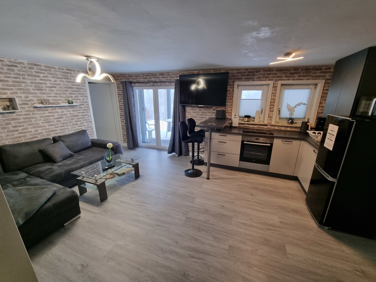 A modern living area features a dark sectional sofa paired with a glass coffee table. A mounted flat-screen TV is positioned above a sleek kitchen area equipped with appliances including an oven and refrigerator. Large sliding doors provide access to an outdoor space, brightening the room.