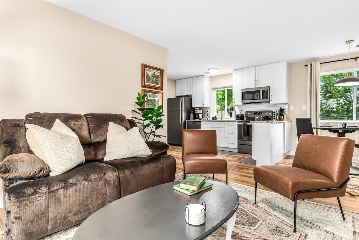 The open living area features a comfortable brown sofa and cozy accent chairs, arranged around a circular coffee table. A kitchen area with modern appliances is visible in the background, complemented by natural light streaming through nearby windows.