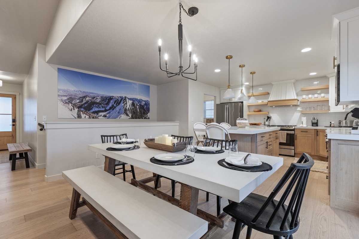 A spacious dining area features a long, rustic table surrounded by black chairs. Modern light fixtures hang above, illuminating the space. The open layout connects to a well-equipped kitchen, showcasing mountain views through a large mural.