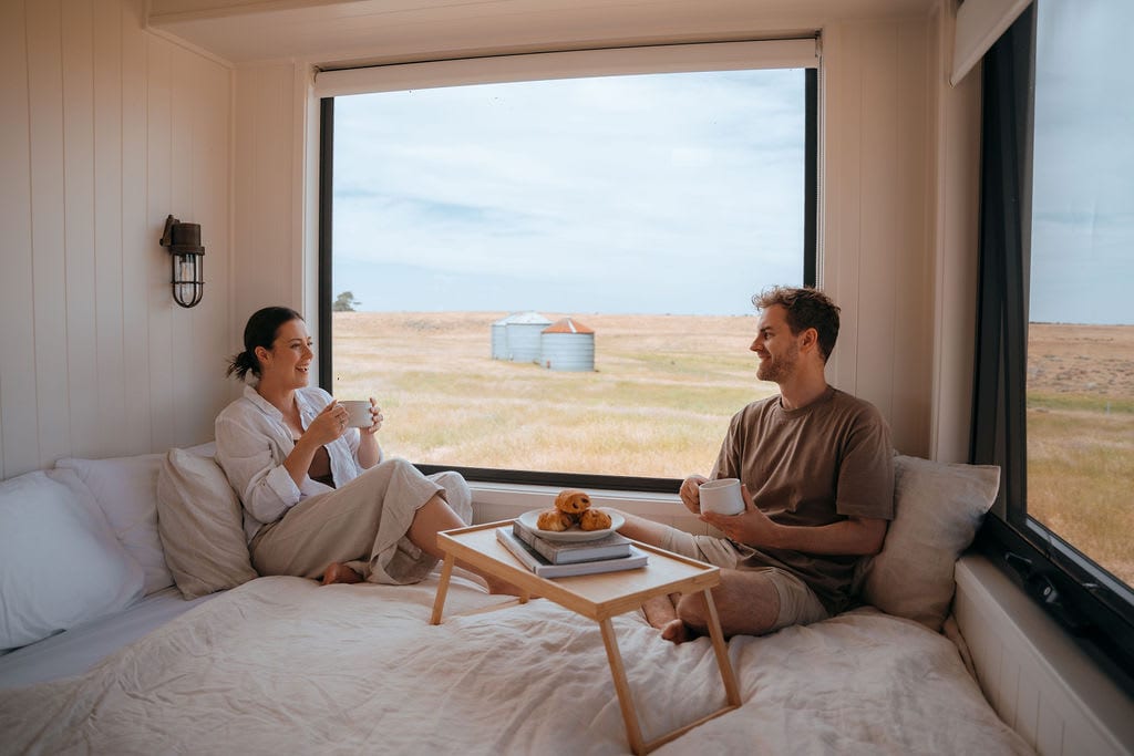 A cozy sleeping area features a spacious bed with soft bedding, complemented by a small tray holding pastries and cups. Large windows frame views of the serene countryside, showcasing distant silos against golden grassy fields, inviting relaxation.