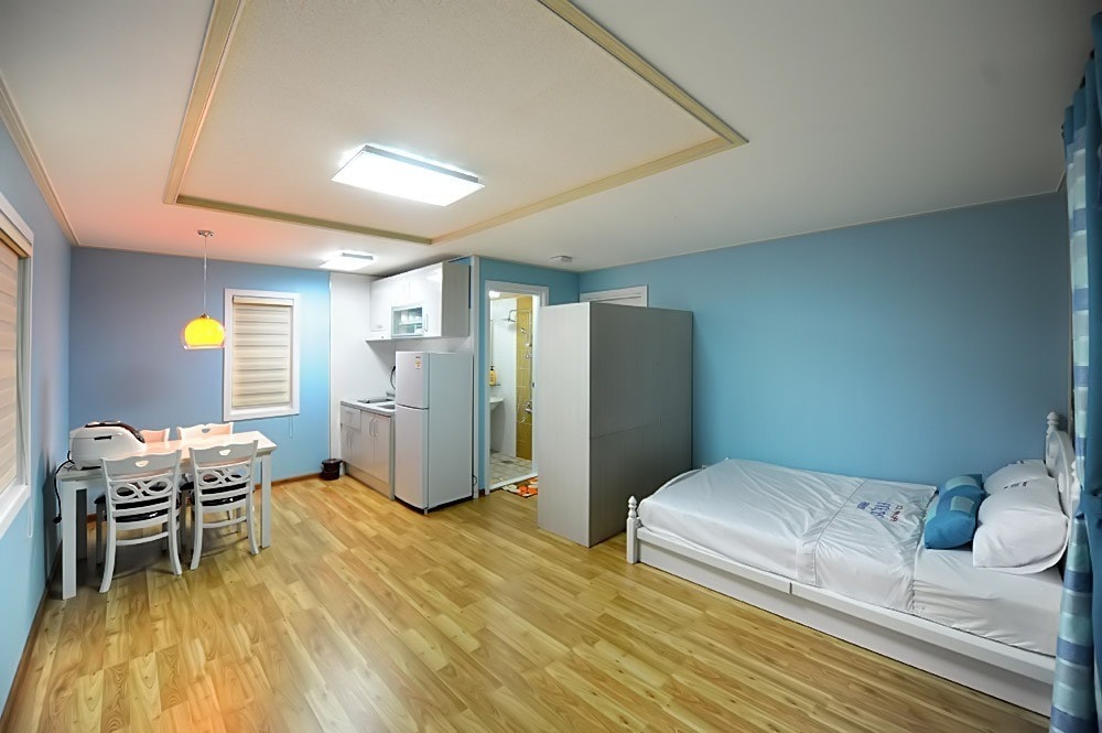 A spacious interior features light blue walls and wooden flooring. A cozy sleeping area is visible, showcasing a spacious bed with white bedding. A dining table with four chairs is arranged nearby, with an open kitchen area in the background.