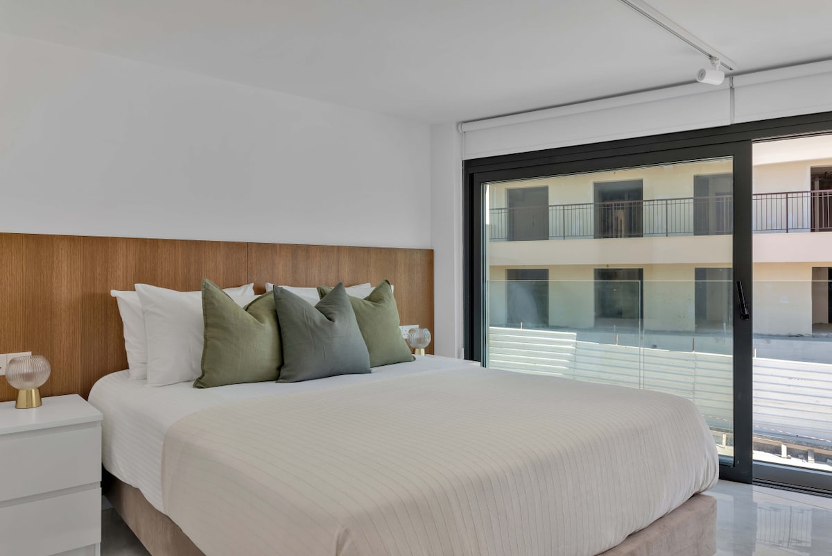 A spacious bedroom features a large queen-size bed adorned with soft white linens and decorative pillows in shades of green. A sleek bedside table with a lamp accompanies the bed. Large glass doors provide natural light and a view of the outdoor area beyond.