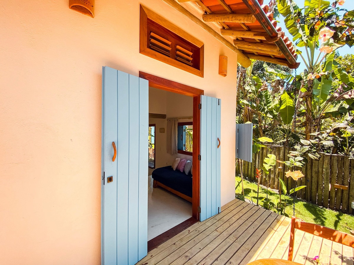 The exterior view shows a charming entryway with open blue shutters, revealing a cozy sleeping area inside. A wooden deck extends outward, surrounded by vibrant greenery. The space includes a dark bed adorned with pink pillows, inviting relaxation.