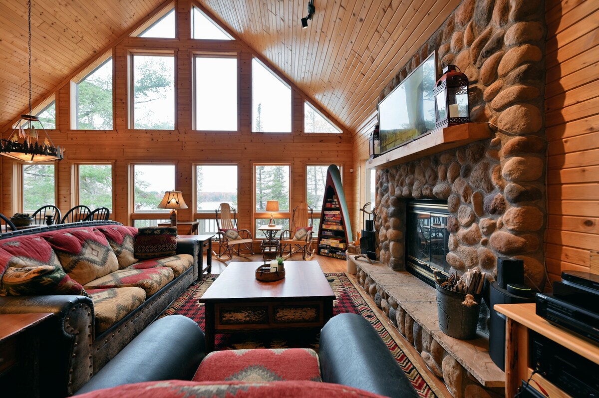 The spacious living area features a stone fireplace as the focal point, surrounded by comfortable seating. Large windows allow natural light to flood the room, highlighting the wooden walls and ceiling. A rustic coffee table sits in the center, complemented by decorative accents.