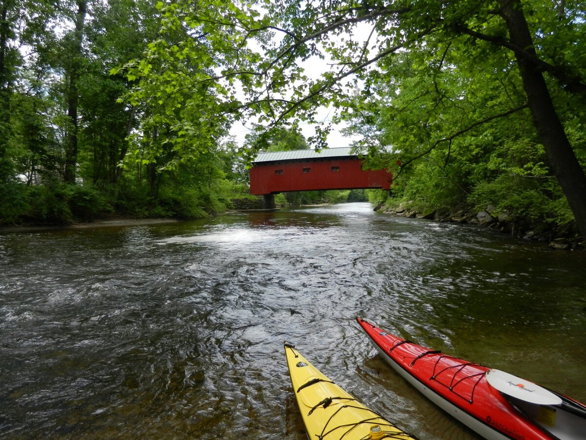 A serene river scene is captured, featuring two kayaks in vibrant yellow and red resting on the water's edge. A charming red covered bridge is visible in the background, surrounded by lush green trees reflecting in the water, creating a peaceful natural setting.
