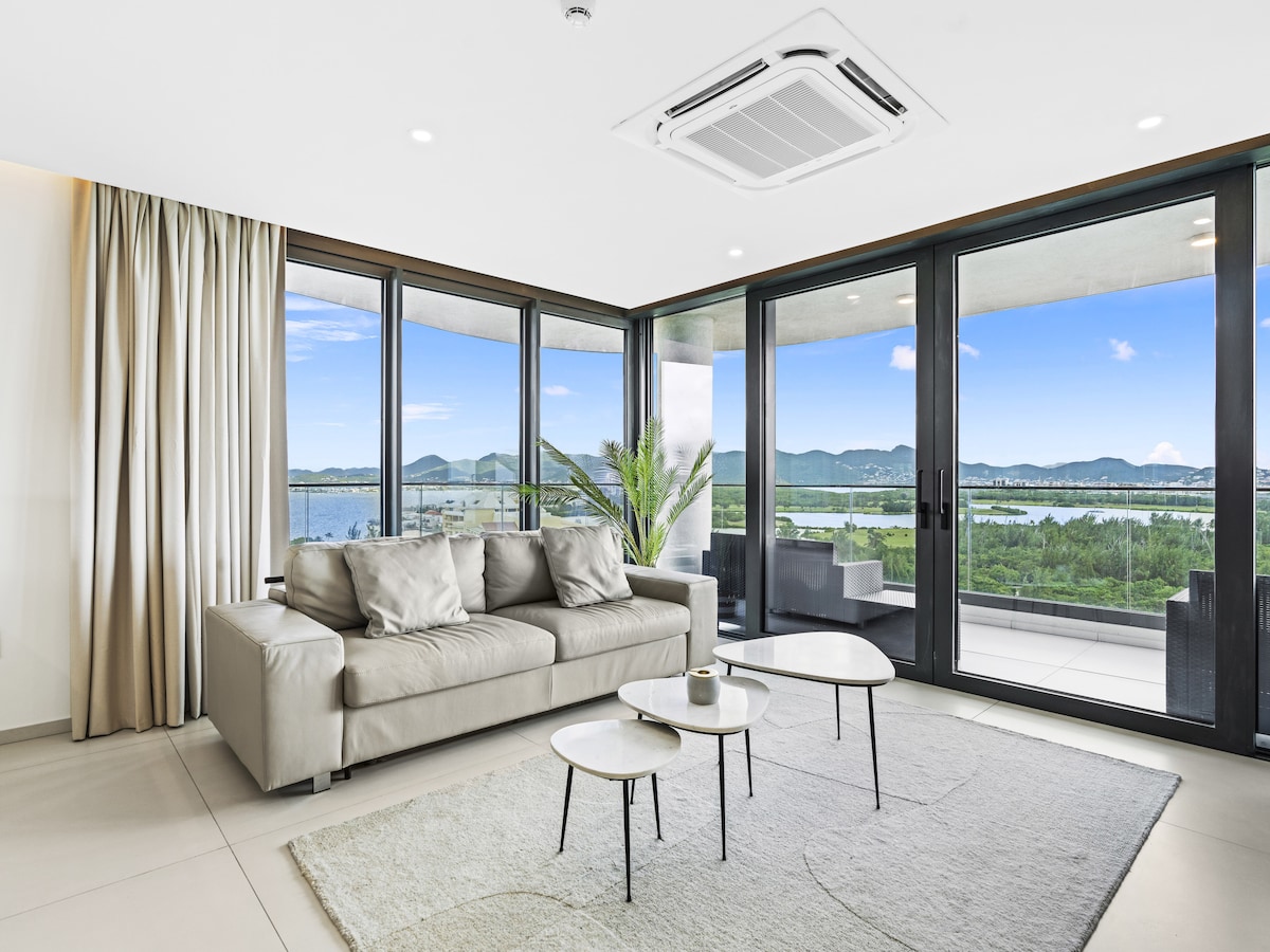 A modern living area features a light-colored sofa facing large glass doors that frame a scenic view of the outdoor landscape. A neutral area rug lies under a set of small tables. Natural light fills the space, complemented by soft curtains.
