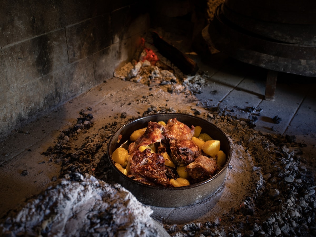 A dish of roasted chicken is placed in a round pan, surrounded by an arrangement of golden potatoes. The meal rests on a bed of ash and charcoal, hinting at a rustic cooking method. Dim light enhances the warm tones of the food.