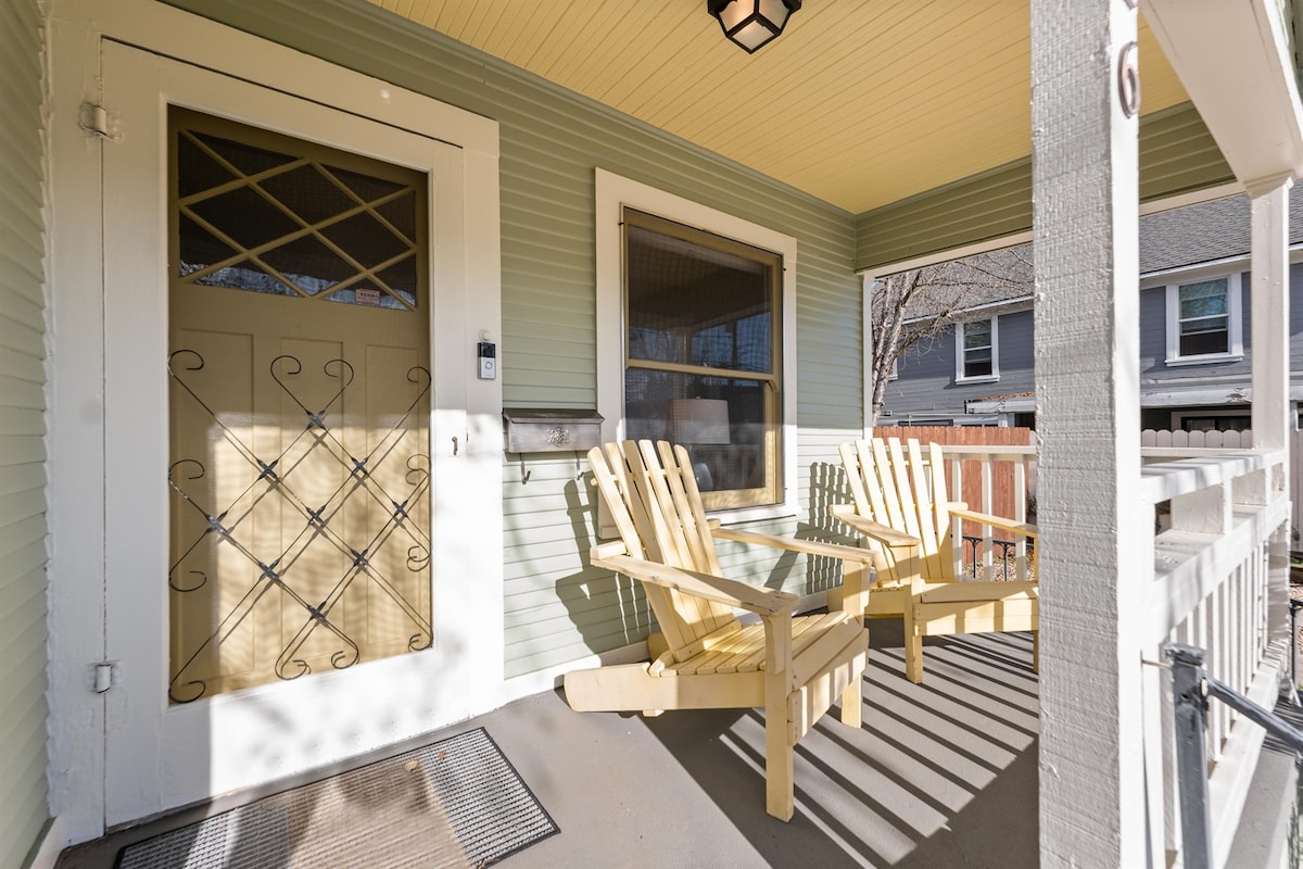 A charming porch is equipped with two wooden chairs, positioned to invite relaxation. Sunshine casts playful shadows through the railing, highlighting the light green walls. The front door features decorative paneling, adding character to the space.