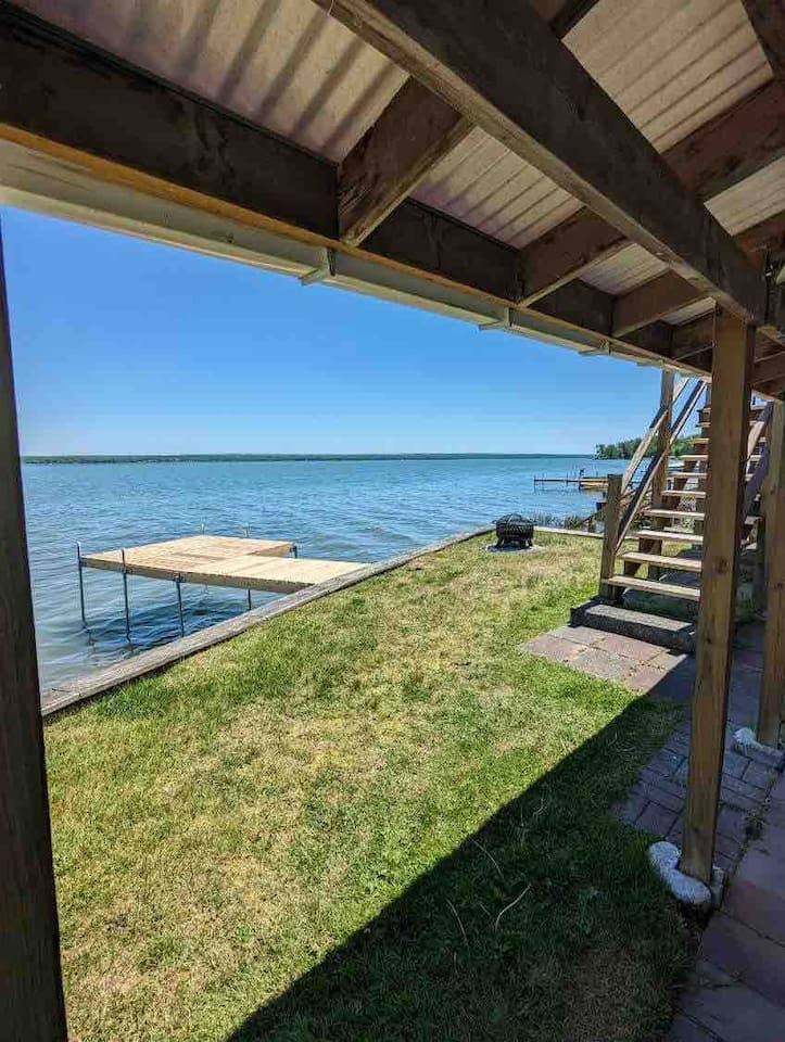 A view of the lakefront reveals a grassy area leading to the water's edge. A wooden dock extends into the lake, and a small grill is positioned nearby. Stairs ascend on the right, providing access to an upper level.