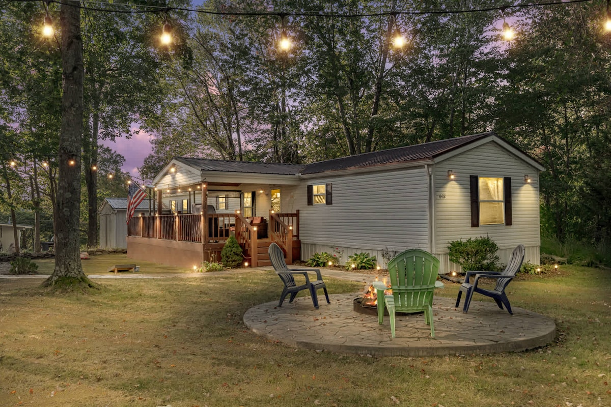 The exterior of the home is illuminated by string lights, creating a cozy ambiance. A fire pit is surrounded by several chairs on a circular stone patio. The home features a deck and is set amidst trees, with a grassy area surrounding it.