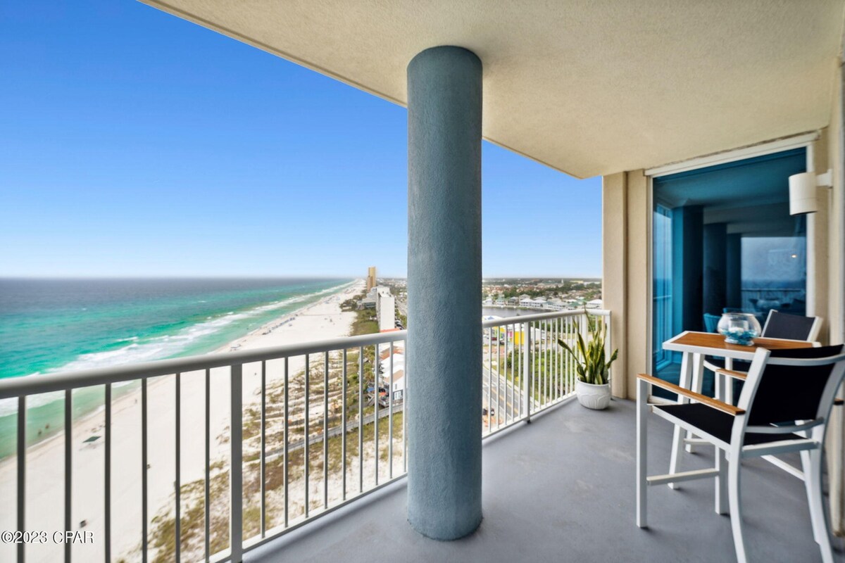 A spacious balcony offers a stunning view of the beach and ocean. The scene is enhanced by a clear sky and gentle waves. Seating is provided by a small table and two chairs, accompanied by a potted plant for a touch of greenery.