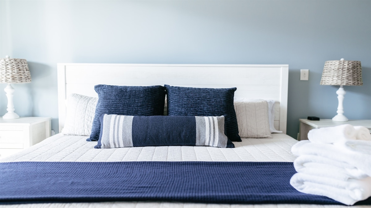 A neatly made bed with navy and white accents draws attention, featuring textured pillows and a long bolster pillow. Soft white towels are neatly stacked at the foot of the bed, while bedside lamps provide a touch of charm to the tranquil setting.