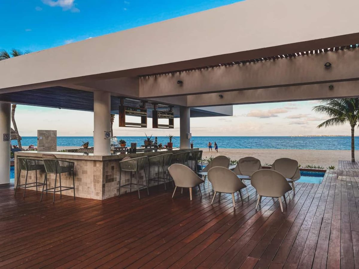 An open-air bar area features a sleek countertop with seating options at the front. Soft, modern chairs are arranged around the space, providing a view of the turquoise sea and sky, creating a relaxed atmosphere for gatherings.