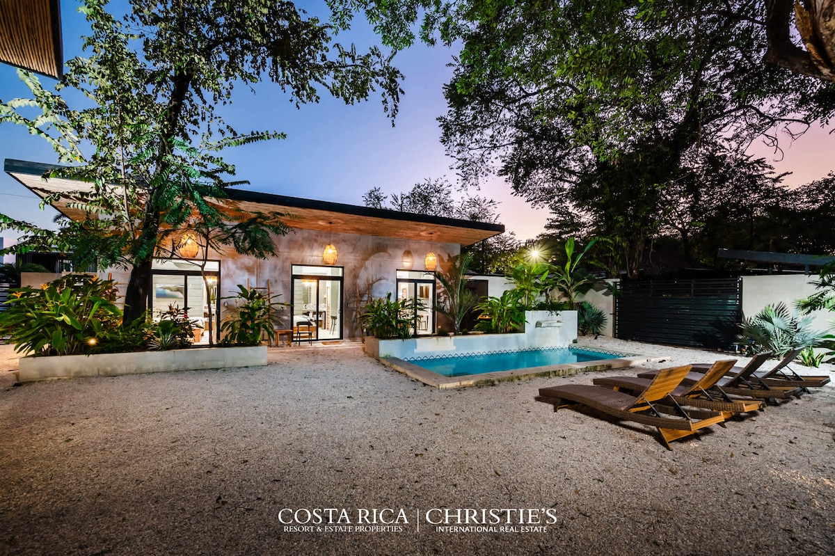 A modern house is surrounded by lush greenery, featuring large glass doors that open to a central pool area. Sun loungers are positioned alongside the pool, and warm lighting highlights the exterior structure against a twilight sky.