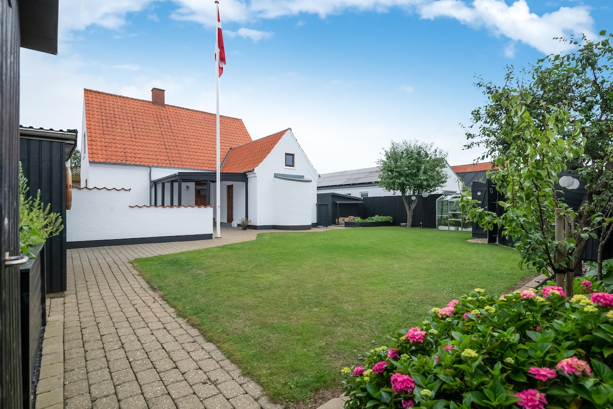 A well-maintained outdoor area features a large lawn surrounded by green foliage and colorful flowering plants. The white exterior of the house is complemented by a red roof and a flag pole. The clean pathway leads to the entrance, providing a welcoming approach.
