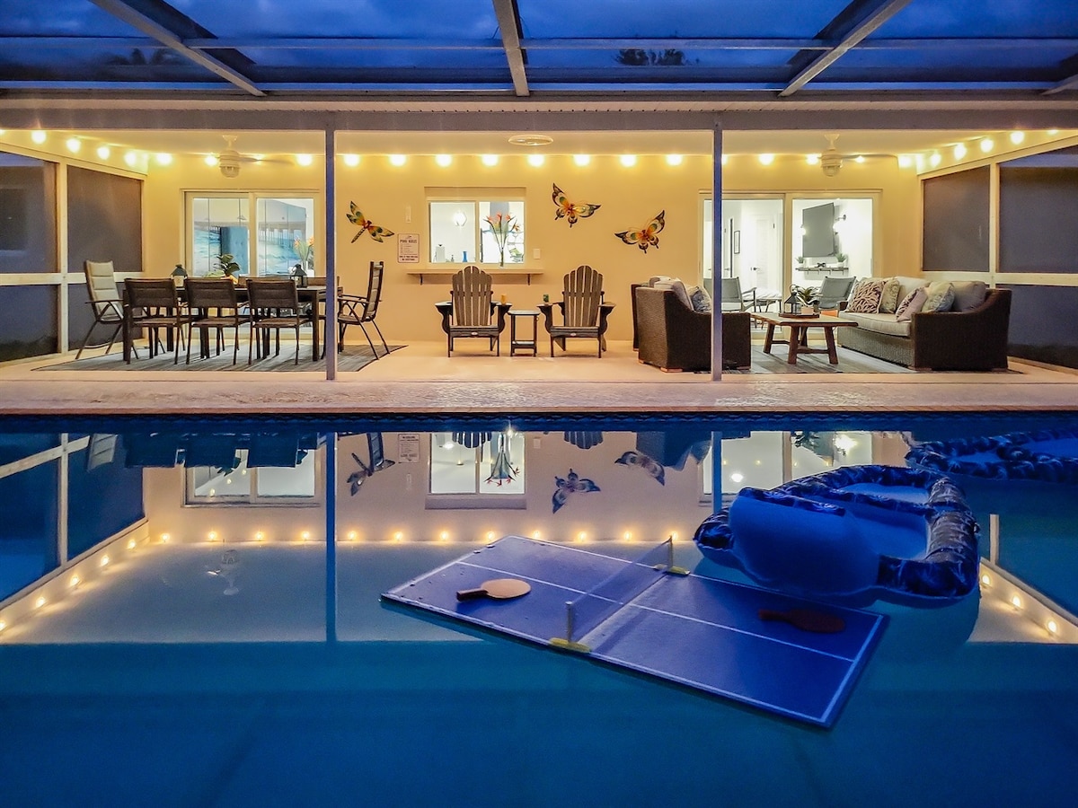 A covered outdoor area is visible, featuring multiple seating arrangements and a dining table under soft lighting. The calm pool reflects the illuminated space, with a pool float and Infinity Table in view, suggesting a welcoming environment for relaxation and entertainment.