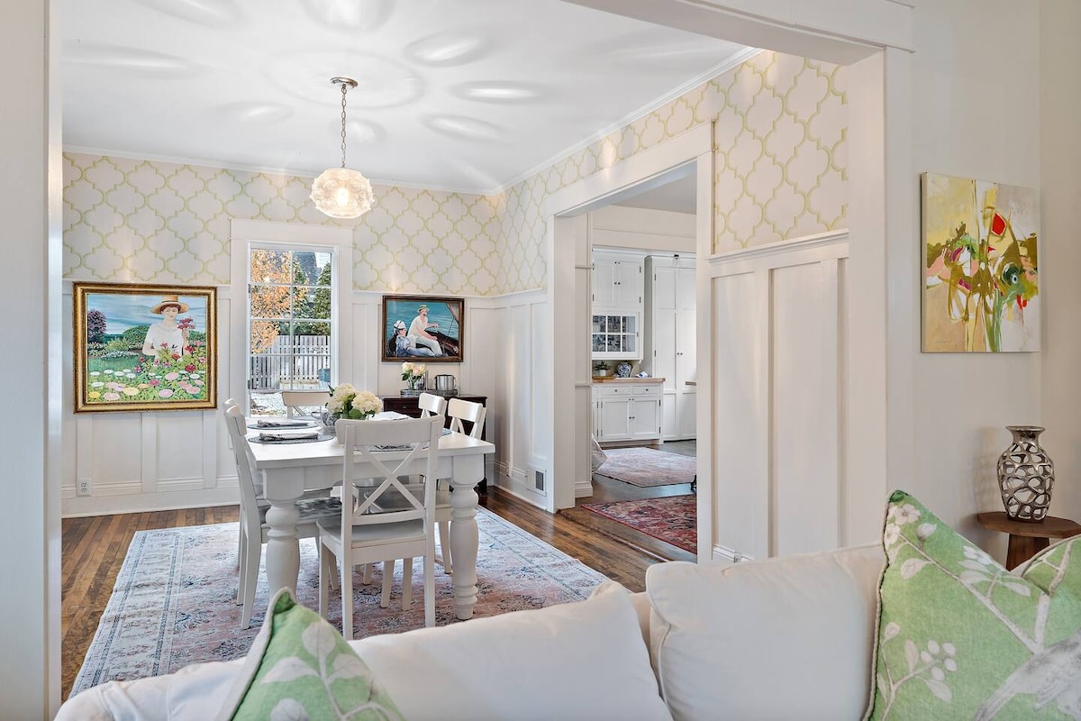 An elegant dining area features a white table surrounded by matching chairs, accented by a decorative chandelier. The walls are adorned with light-colored wallpaper and framed artwork, while a view into the kitchen is visible, highlighting an inviting and airy atmosphere.
