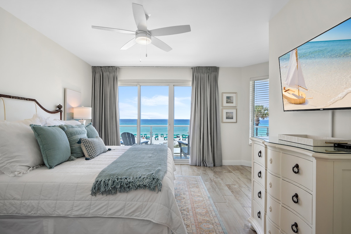 A spacious bedroom features a large bed adorned with soft pillows and a decorative blanket. Natural light highlights the room, which opens to a private balcony offering expansive views of the beach and ocean. A ceiling fan and a television are also present.