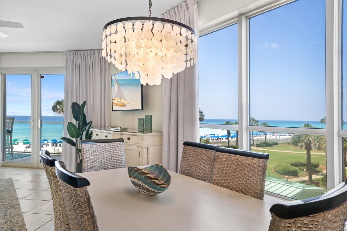 A dining area is highlighted by a chandelier featuring circular accents above a light-colored table. Woven chairs surround the table, and a decorative bowl rests in the center. Large windows provide a view of the beach and ocean, enhancing the bright atmosphere.