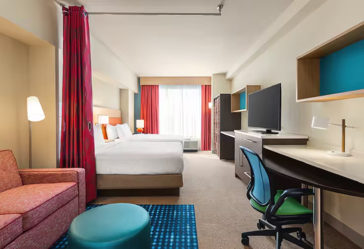 The studio suite features two queen beds with crisp white linens, complemented by a cozy sitting area with a vibrant pink chair and a turquoise ottoman. A large window allows natural light to brighten the space, while a desk and modern television provide convenient amenities.