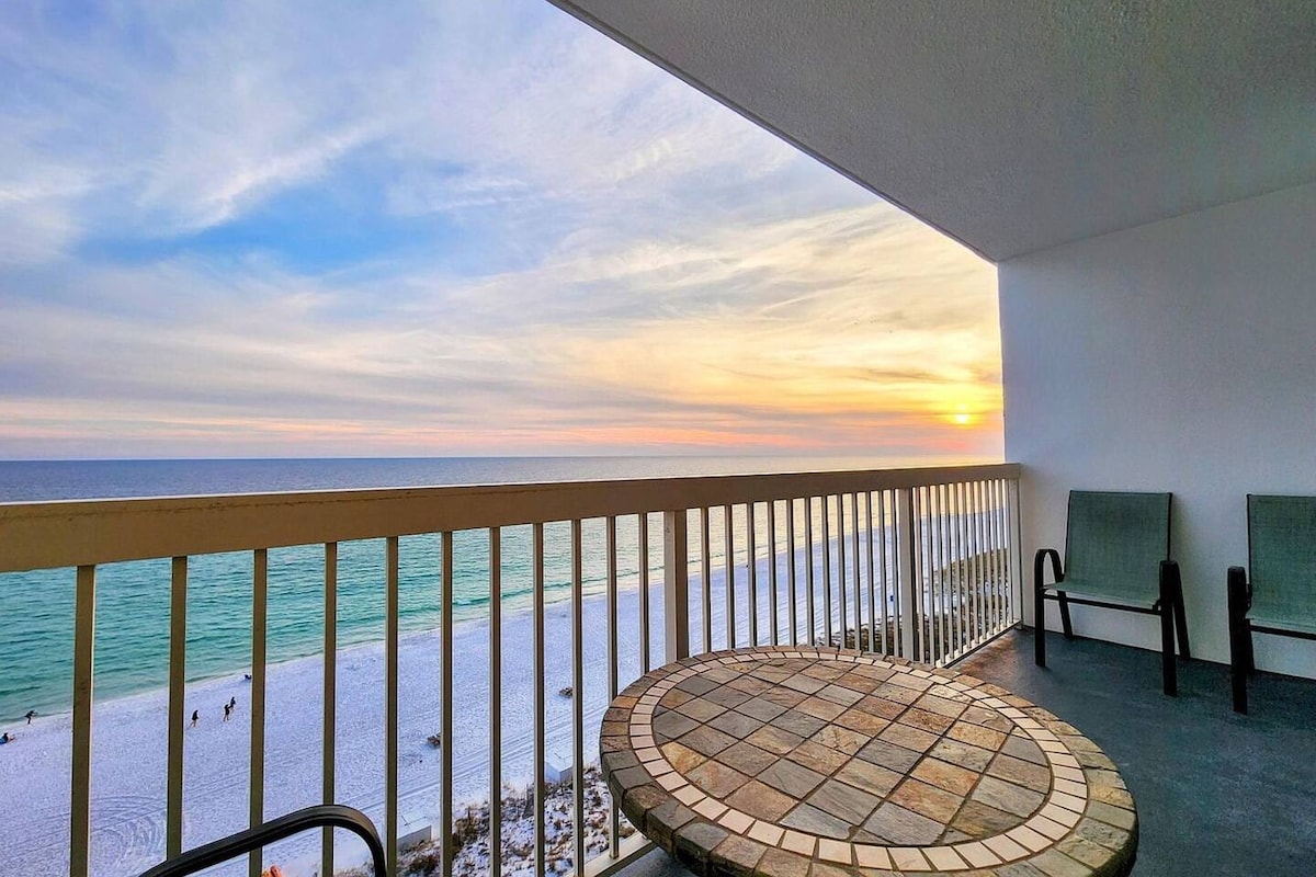 A private balcony offers a stunning view of the Gulf of Mexico, with the sun setting over the horizon. Two chairs are positioned beside a circular table, creating a comfortable space to enjoy the ocean scenery. The white sandy beach is visible below.