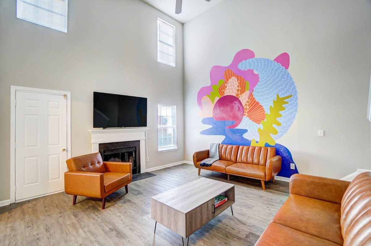 An airy living room is highlighted by a large mural, featuring vibrant colors and abstract shapes. Two leather sofas and a modern coffee table are arranged around a fireplace. Natural light filters through large windows, enhancing the spacious ambiance.