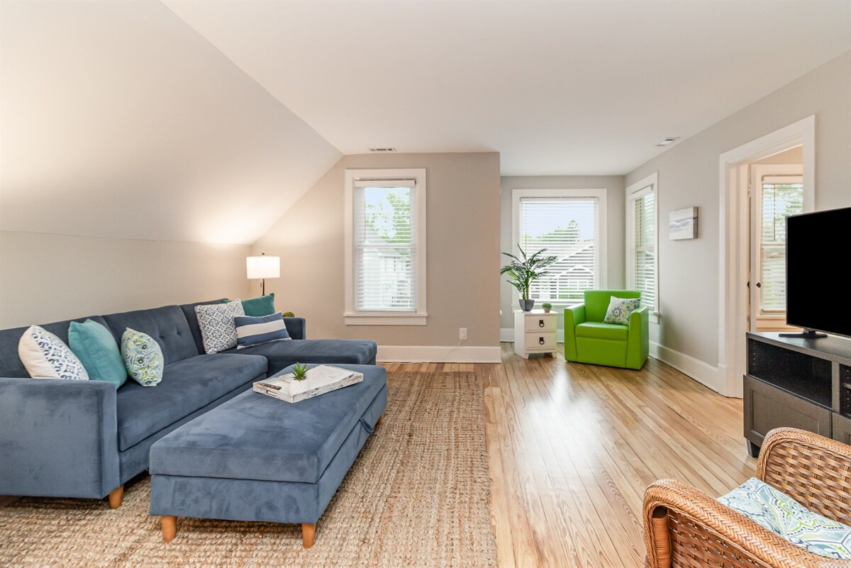A spacious living room features a large blue sectional sofa with decorative cushions and a woven area rug. Natural light floods the space through multiple windows, illuminating the neutral wall color. A green accent chair and a small plant add a touch of color, while a TV is positioned nearby.