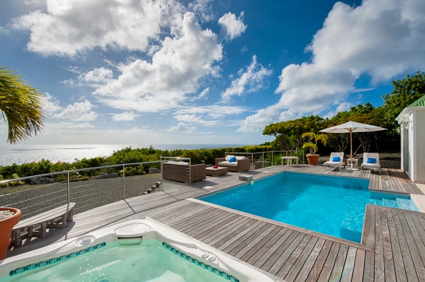 An outdoor terrace features a swimming pool and a jacuzzi, surrounded by wooden decking. Recliners are positioned by the pool, while an umbrella provides shade nearby. Lush greenery frames the scene, with ocean views extending to the horizon under a partly cloudy sky.