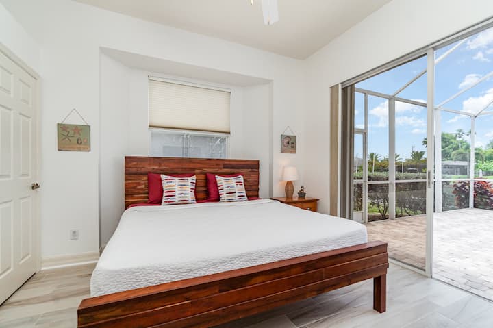 Dhomë gjumi e ajrosur me një kornizë moderne krevati prej druri, shtroja të bardha të freskëta dhe qasje të drejtpërdrejtë në një verandë të ndriçuar nga dielli.