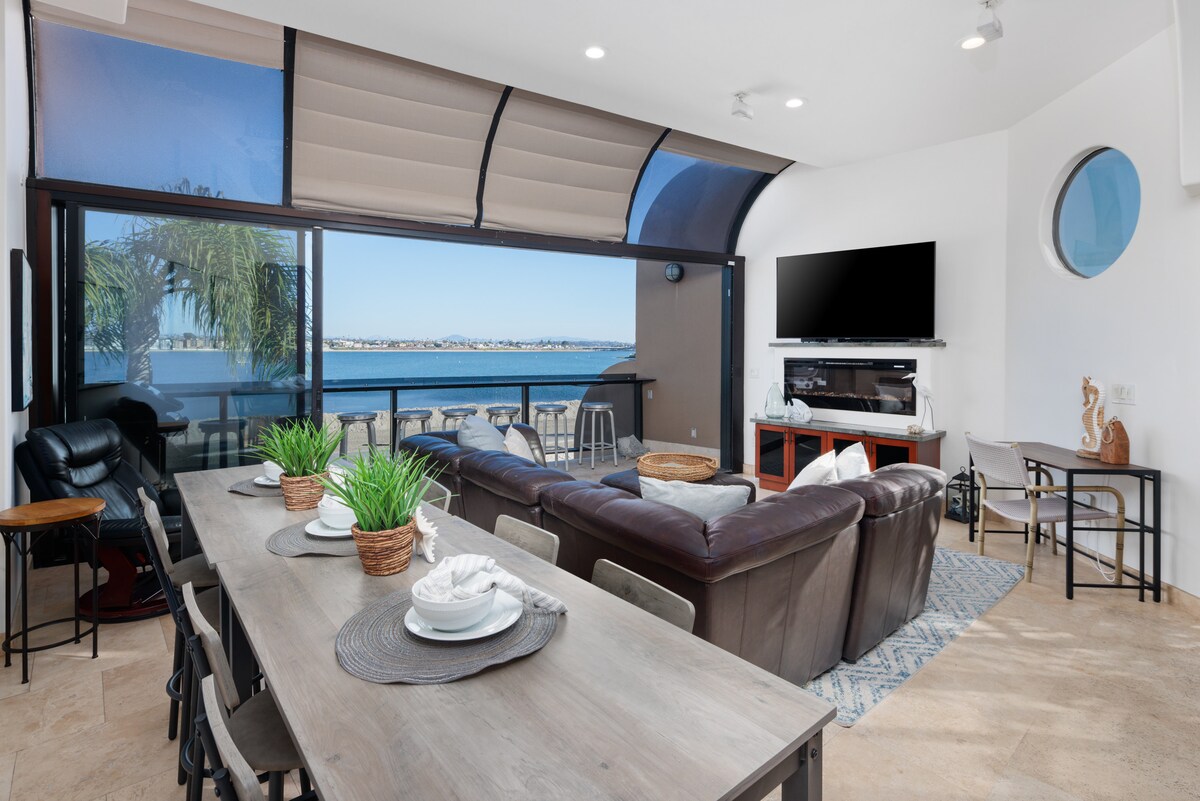 A spacious living area features large glass sliding doors that open to stunning water views. Comfortable seating is arranged around a coffee table, with a dining table nearby. A flat-screen television is mounted on the wall, and decorative elements add a coastal touch.