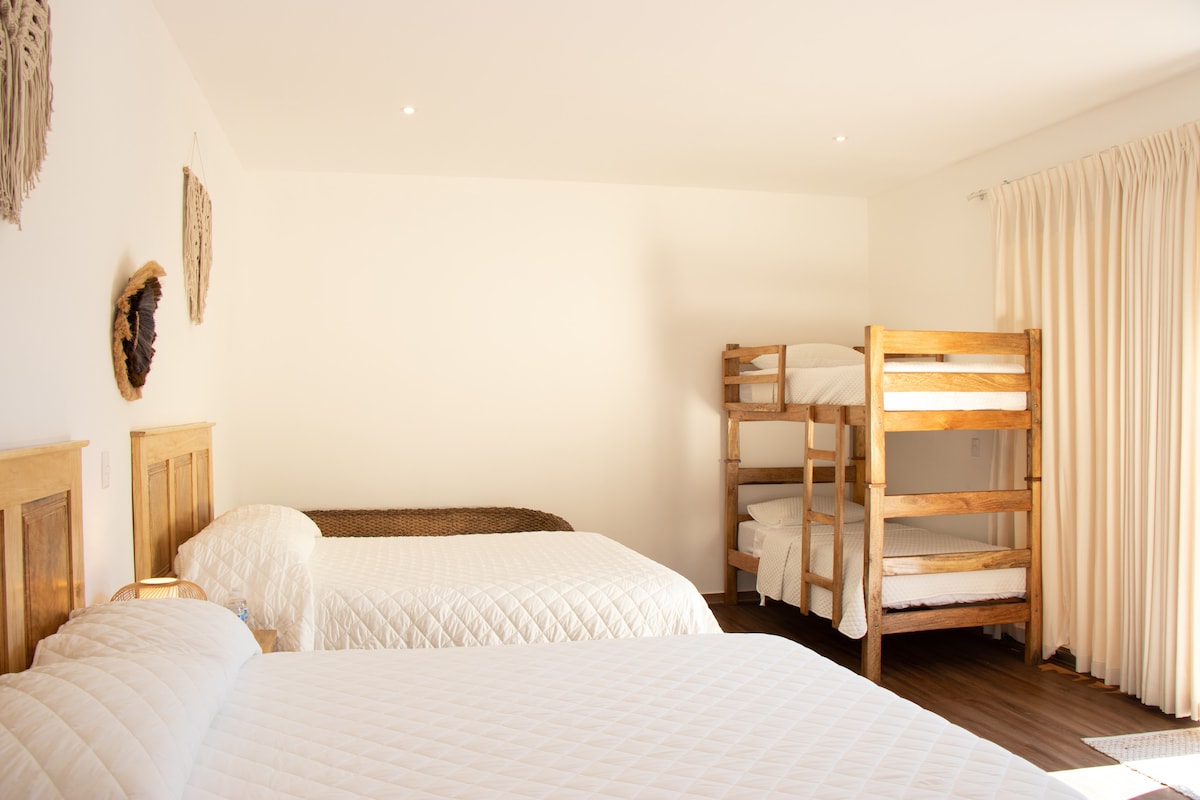 A spacious family room features two double beds and a wooden bunk bed, creating comfortable sleeping arrangements. Natural light filters through large windows with light curtains, illuminating the hardwood floor. Minimalist decor adds a serene touch to the inviting space.