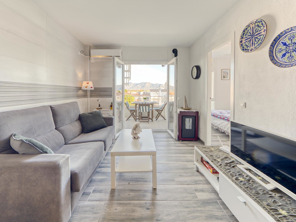 A modern living area features a comfortable gray sofa and a white coffee table. Natural light flows in through the glass doors that open to a balcony, showcasing views of the surrounding mountains. Decorative plates adorn the walls, adding a touch of character.