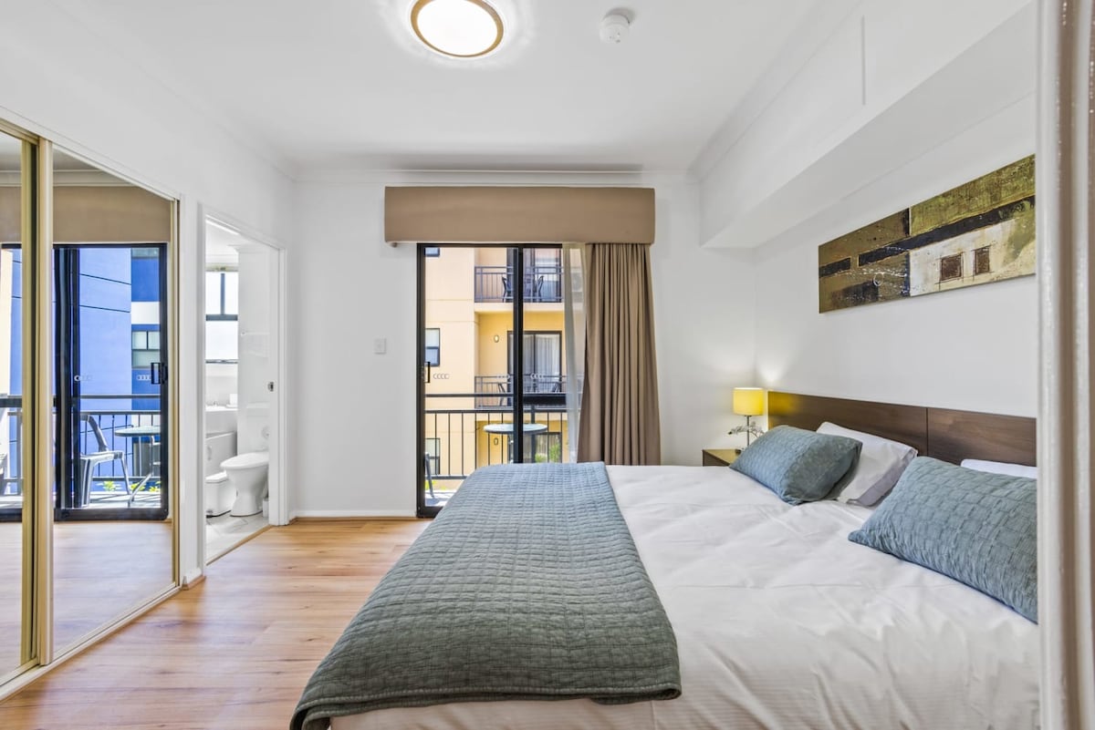 The bedroom features a spacious king-sized bed adorned with soft linens and throw pillows. Natural light filters through the balcony doors, illuminating tasteful wall art. A mirrored wardrobe adds a contemporary touch, while an ensuite bathroom is visible through an open door.