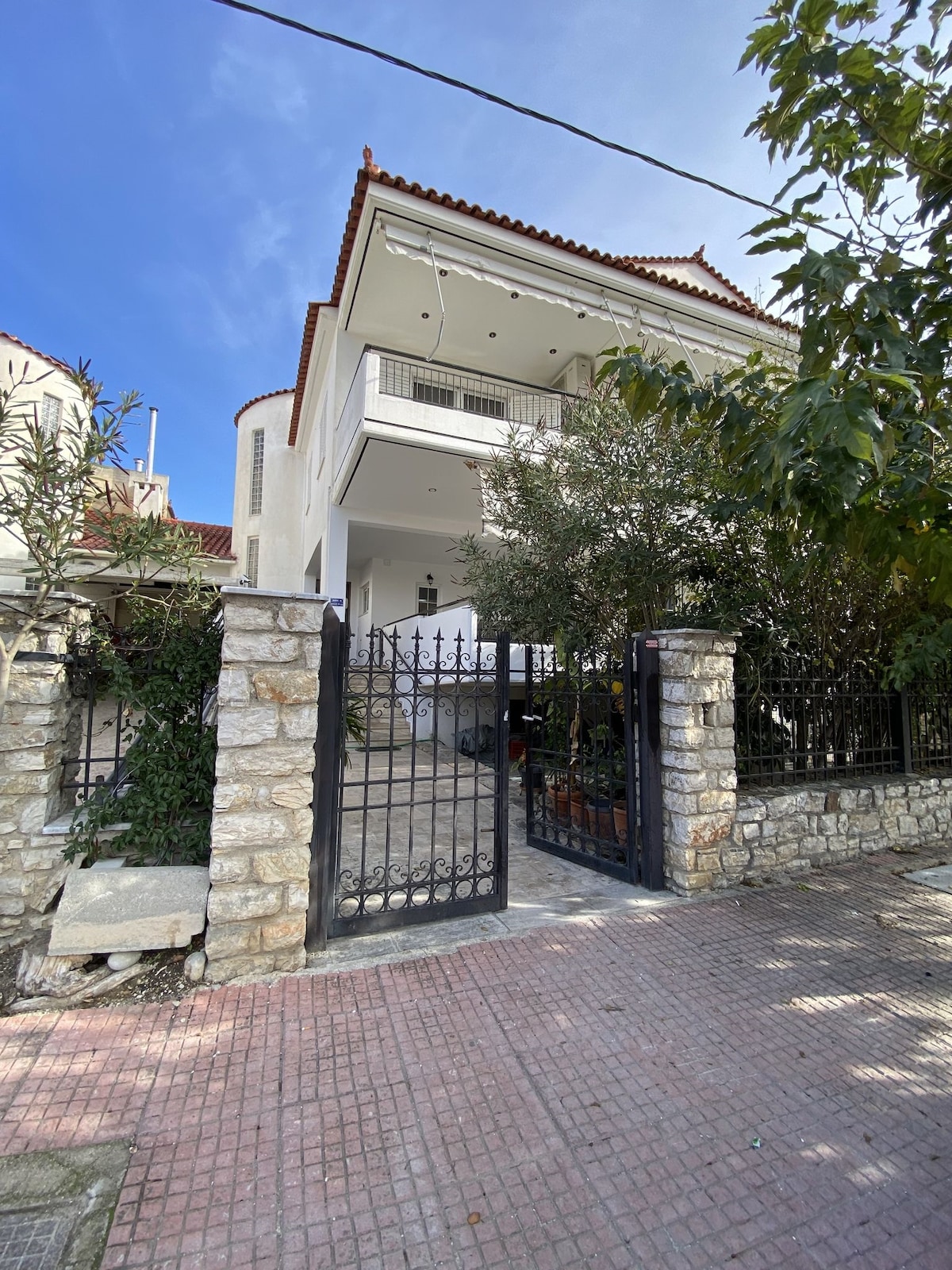 The exterior of the apartment is shown, featuring a two-story building with a red-tiled roof. A stone wall surrounds the property, complemented by a wrought-iron gate. Lush greenery is visible near the entrance, and the clear blue sky adds to the welcoming atmosphere.