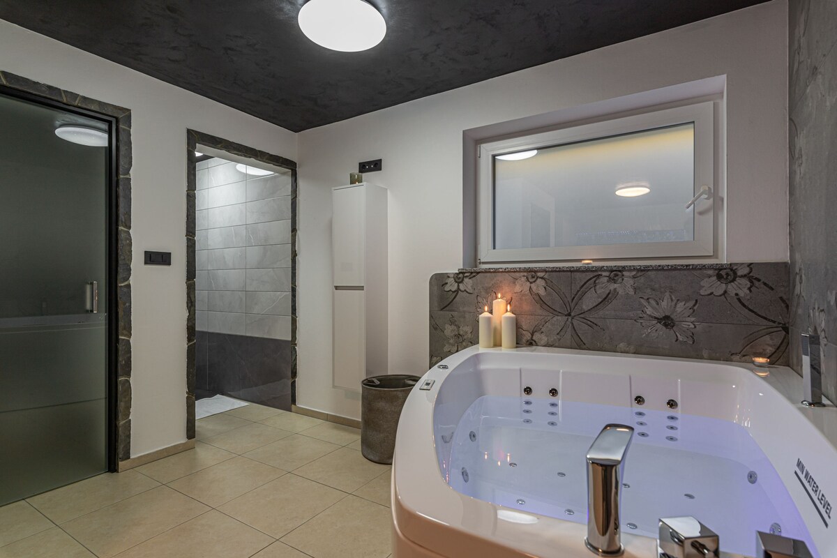 An ensuite bathroom features a spacious massage tub for two, with soft lighting provided by candles nearby. The room includes a glass door leading to a shower area, complemented by light-colored tiles and floral accents on the walls.