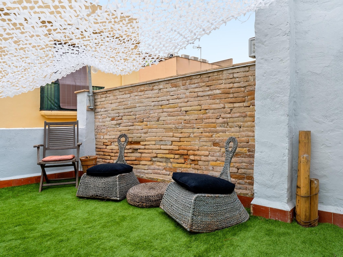 An outdoor terrace features a grassy area with two woven seating options adorned with black cushions. A low round table is positioned between the seating. A translucent canopy drapes overhead, providing shade and creating a relaxed atmosphere against a backdrop of a textured stone wall.