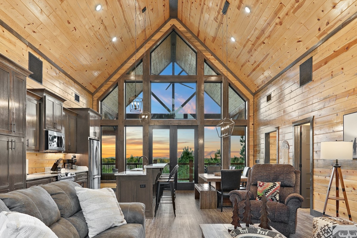 A spacious living area is highlighted by towering windows that frame picturesque mountain views and vibrant sunsets. Wooden interiors enhance the cozy atmosphere, complemented by comfortable seating and a stylish dining area. Modern appliances in the kitchen can be seen, creating a welcoming space for gathering.