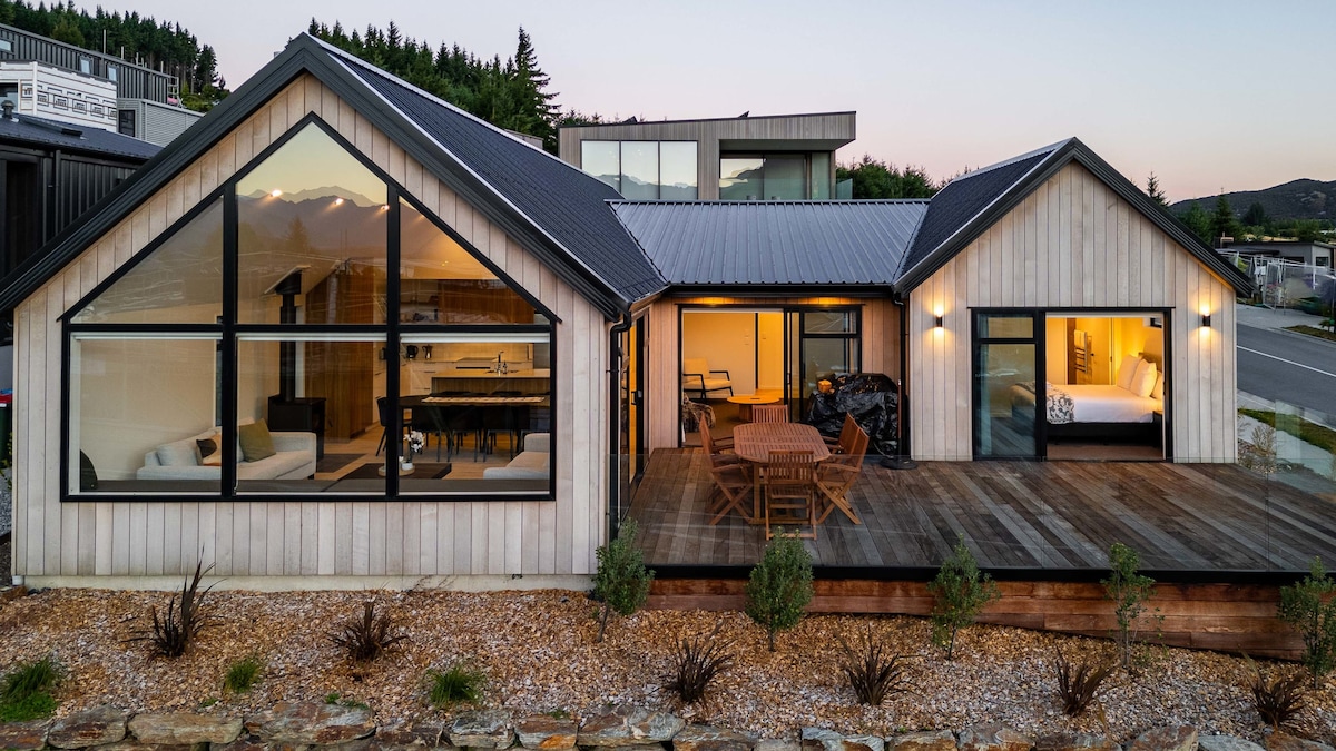 A modern architectural design showcases a combination of cedar cladding and large windows. An expansive deck is furnished with a wooden dining table and chairs, providing a welcoming outdoor space. Soft lighting illuminates the interior areas, hinting at the cozy atmosphere within.