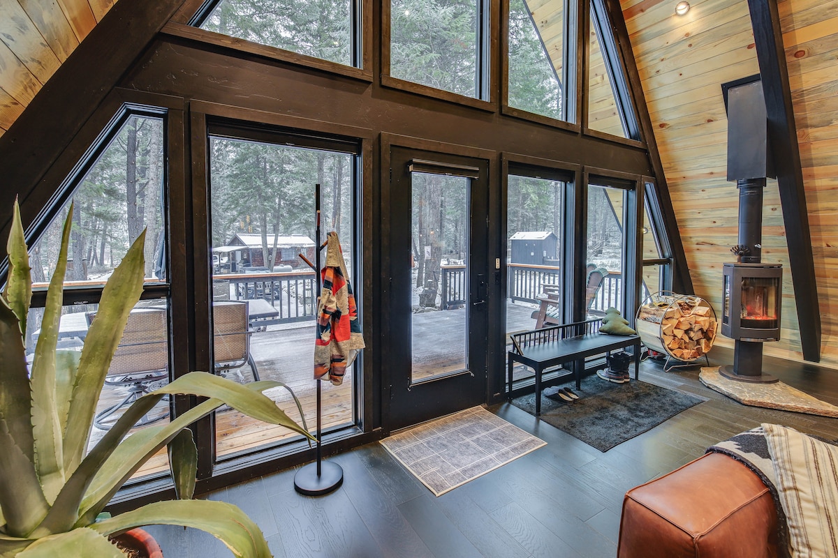 The interior space features a striking A-frame design with floor-to-ceiling windows framing views of the outdoor wooded area. A cozy wood-burning stove stands nearby, and a potted plant adds a touch of greenery. Natural light fills the room, creating a warm environment.
