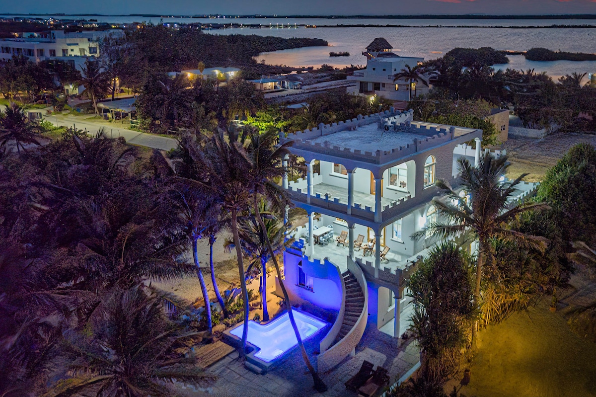 An aerial view of a three-story home is presented, featuring a private swimming pool surrounded by lush tropical landscaping. The exterior is illuminated, showcasing the spacious balconies and large windows. The ocean can be seen in the background, creating a serene coastal ambiance.