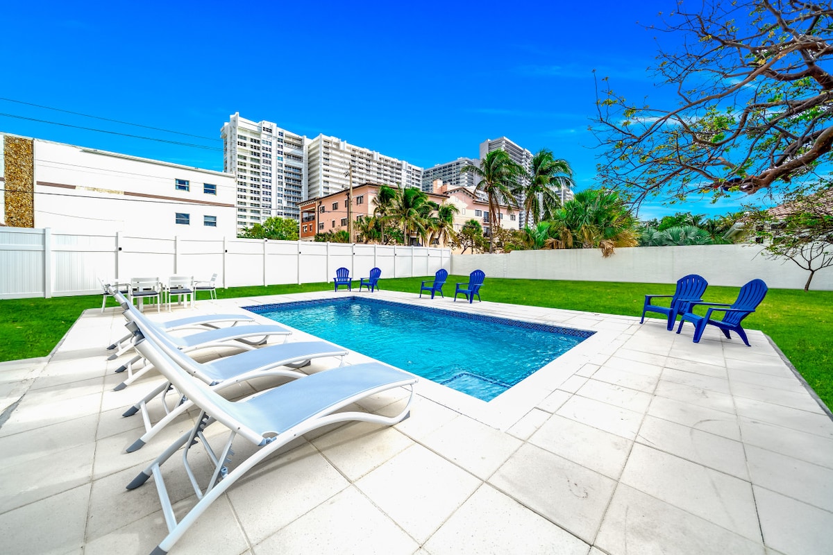 A spacious backyard is highlighted by a large heated pool surrounded by lounge chairs and stationary seating. Lush greenery and palm trees are visible, providing a tropical atmosphere. The backdrop features several tall buildings, emphasizing the property's proximity to urban amenities.