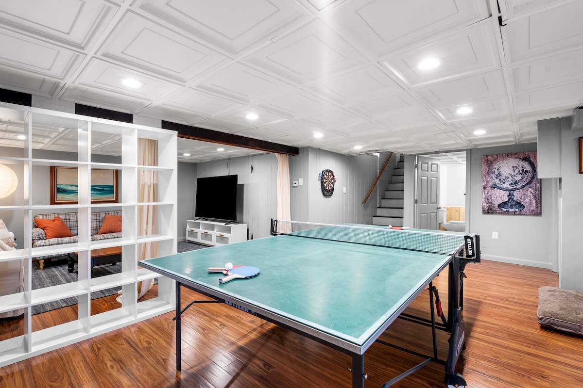 A spacious basement area features a ping pong table at the center, surrounded by seating and a smart TV. Soft lighting enhances the room's warm ambiance, with a decorative wall art piece visible in the background. Stairs provide access to the main level.