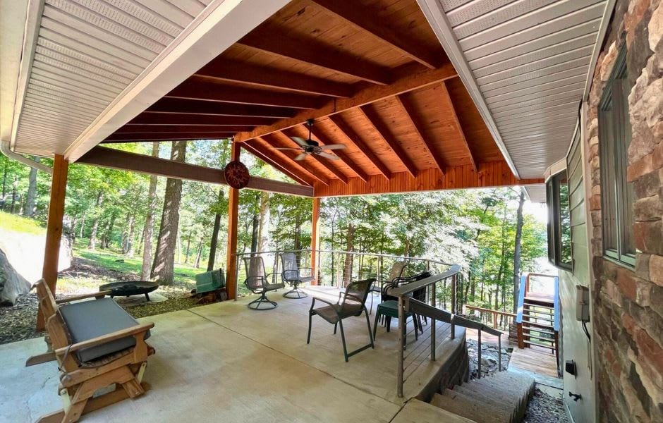 A spacious covered deck features a wooden ceiling and ample seating options, including a sofa and dining set. A ceiling fan enhances comfort, while surrounding trees create a natural backdrop. Steps lead to additional outdoor areas, providing a peaceful space for relaxation.