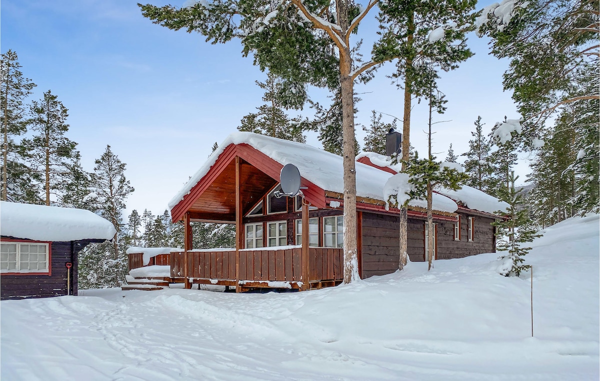 A charming vacation home is set against a snowy landscape, showcasing a sloped roof with snow accumulation. The exterior features wooden walls and a spacious deck, offering an inviting entrance. Tall trees surround the area, emphasizing the peaceful, winter ambiance of the location.
